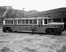 A long, 8-wheeled city bus is viewed from the side, parked in front of a brick building. The bus is about 1.5 times as tall as the uniformed man in the foreground.