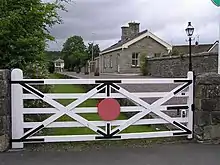 An ornate white gate obscures the view of a small stone station building and platform, with grass growing where the railway tracks would have been. In the distance at the far end of the platform is the original signal building.