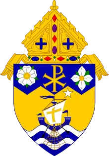 Shield topped by a mitre, featuring a white heraldic rose, Chi Rho, and Pacific dogwood on blue field at top; the Barque of St. Peter and a white star on gold field; and four alternating blue and white wavy stripes at the bottom