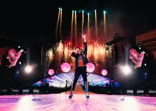 A man performs with a microphone as the stage behind him release numerous fireworks