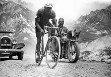 A cyclist climbing a mountain, followed by a motorcycle and a car.