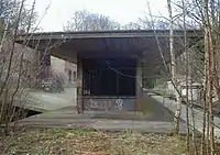 A brick building missing windows, projecting canopy and graffiti. Tree saplings and vegetation grow from the platform surface.