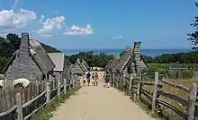 A modern-day photograph of a village consisting of small, primitive wooden houses. Most of the houses have thatched roofs. In the distance is a large expanse of ocean and a clear blue sky.