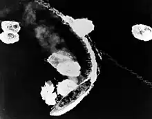 An overhead view of a large warship partway through a turn to the right. The ship's wake curves around behind her, and the surrounding sea is dotted with large areas of disturbed water and foam.