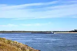 An open stretch of river water. A barge is in the center of the image, and in the background is a wooded riverbank. There are no signs of human habitation on the riverbanks.