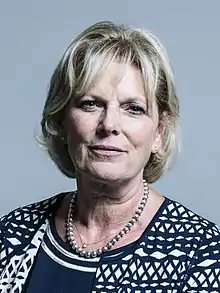 Close-up of a white woman with blonde hair, blue eyes and a neutral facial expression. She is wearing subtle makeup, a dark blue dress with two embroidered bands of white near the neck, a dark blue and white patterned cardigan, a pearl necklace and a thin silver neck chain with a diamond on it.