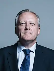 Close-up of a white man with grey hair, blue eyes and a neutral facial expression. He is wearing a blue patterned tie, a light blue and white thin striped shirt and a black suit jacket.