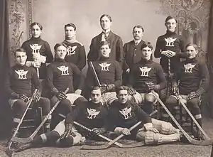 Black and white image twelve men posing for a photo. They are arranged in three rows, five men in the back and middle row, two in the front. The back row is standing, the middle row sitting, and the front row lying on their side towards each other. Ten of the men are wearing a dark wool sweater with an overlapping "P" and "L" with wings, each holding a hockey stick and wearing skates. The other two men are dressed in suits.