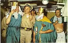 Color photograph of crossing the line ceremony held in the crews mess compartment of the nuclear submarine USS Triton that includes (from left to right) crewman Wilmot A. Jones dressed as the Queen of the Royal Court; Captain Edward L. Beach dressed in khaki uniform and ceremonial sword; Chief Loyd Garlock dressed as King Neptune, Ruler of the Raging Main; crewman Ross MacGregor dressed as Davy Jones in background; and crewman Harry Olsen dressed as the Royal Baby.