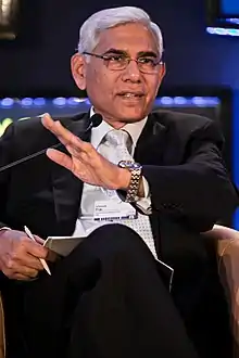 Photograph of a man wearing black suit and left hand in the air.